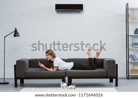 happy young woman using smartphone while lying on sofa under air conditioner hanging on wall