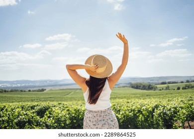 Happy young woman walks through the vineyard. White female in casual clothes and straw hats look at the landscape. Happiness and travel concept. Copy space.