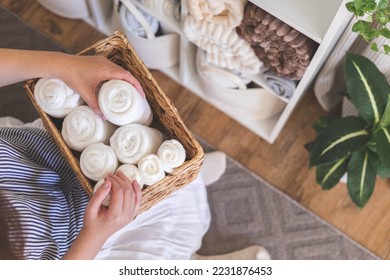 Happy young woman in warm robe is holding stack of rolled organic cotton bath towels in white basket and organizing linen closet with bed sheets, blankets. Nordic style and minimalism. Decluttering.
