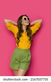 Happy young woman wearing a linen shirt, shorts and sunglasses poses with hands behind head and looks away. Three quarter length studio shot against pink background. - Shutterstock ID 2167037735