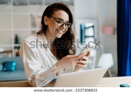 Happy young woman in white shirt and glasses smiling and browsing cellphone while sitting at table with laptop during remote work at home