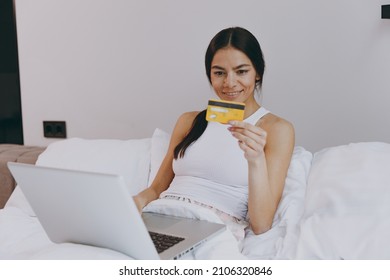 Happy Young Woman In White Tank Top Sit In Bed Using Laptop Pc Computer Holding Looking At Credit Bank Card Doing Online Shopping In Bedroom Lounge Home In Own Room House Wake Up Be Lost In Reverie