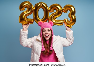 Happy young woman in winter coat holding gold colored numbers against blue background - Shutterstock ID 2220201651
