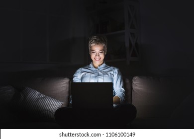 Happy Young Woman Working Late At Home Office, Using Laptop, Copy Space. Casual Girl In Screen Light In Dark Room Background. Technology And Overworking Concept