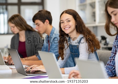 Happy young woman working on laptop and looking at camera in classroom. Portrait of smiling university student in library use computer for a research. Satisfied college student looking at camera.
