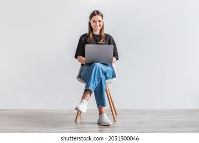 Happy young woman working online, sitting on chair and using laptop against white studio wall, full length. Positive Caucasian lady surfing internet, watching webinar on portable computer