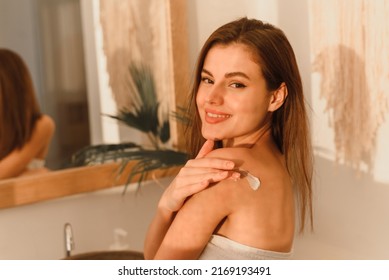 Happy young woman wrapped in towel using nourishing cream or body lotion for her smooth and silky skin after morning shower, rubbing her shoulder, copy space, sun glare. Web banner