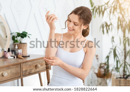 Happy young woman wrapped in towel using body nourishing cream or lotion for her elbows. Pretty lady using moisturizer for her smooth and silky skin after morning shower, copy space, sun flare