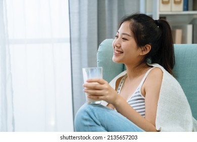Happy young woman wrapped with warm blanket sitting on comfort sofa drinking glass of milk at home living room. charming girl enjoy breakfast beverage in single armchair indoors looking side window