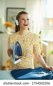 Happy Young Woman In Yellow Dress Ironing On Ironing Board While Listening To The Music With Headphones At Modern Home In Sunny Day.