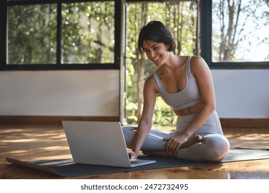 Happy young woman yoga coach trainer sitting on mat in yoga gym studio using laptop computer. Smiling fit Hispanic lady wearing sportswear learning virtual remote online yoga classes on pc. - Powered by Shutterstock