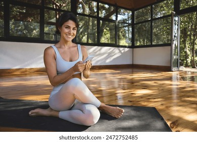 Happy young woman yoga coach sitting on mat in yoga gym studio using smartphone. Smiling fit Hispanic lady wearing sportswear learning sport training exercises in mobile app on cell phone. Portrait. - Powered by Shutterstock