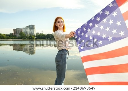 Happy young womanl with USA national flag on her shoulders with high city buildings in background celebrating United States independence day. International day of democracy concept.