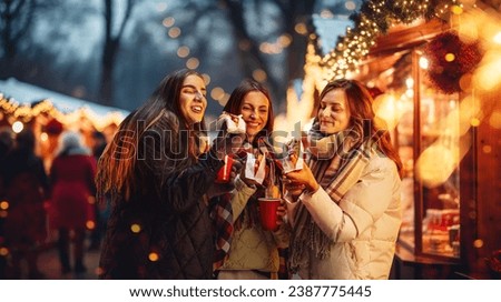 Happy young women attending winter fair in evening, drinking hot drinks, eating hot dogs. Merry season and cheerful time. Concept of winter holidays, Christmas, traditions, outdoor fair, happiness