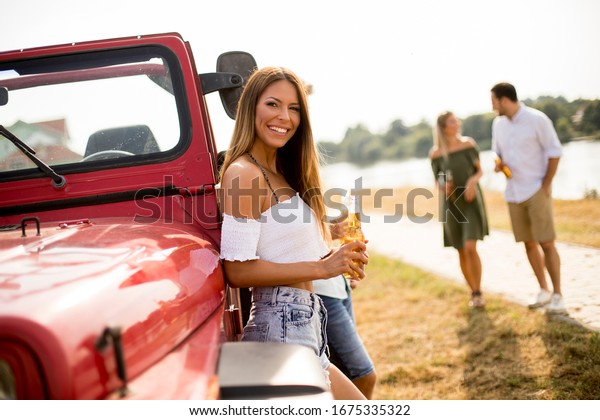 Happy young women drinks cider from the\
bottle by the convertible car with\
friends