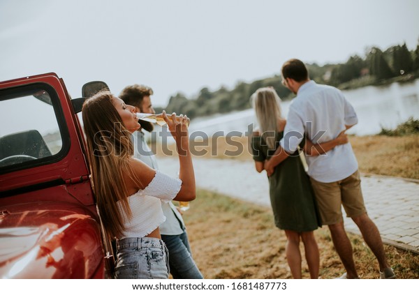 Happy young women drinks cider from the\
bottle by the convertible car with\
friends
