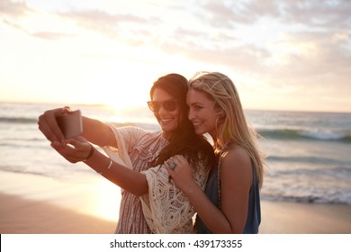 Happy Young Women Enjoying Vacation Together Having Fun On The Beach And Taking Selfie Photo Using Smartphone Camera.