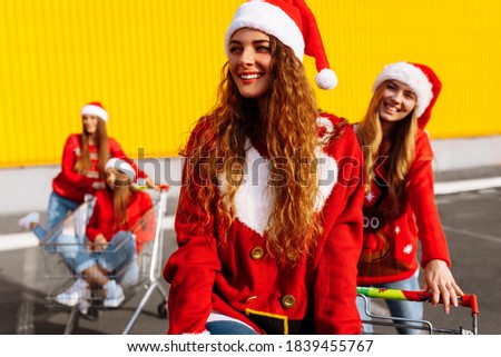 Happy young women friends in christmas sweaters and santa claus hats celebrate new year and ride trolleys against yellow wall background