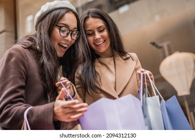 Happy young women looking into shopping bags of her friend near shop showcase outdoor. Buying clothes presents for holidays on sale discount. Black friday - Shutterstock ID 2094312130