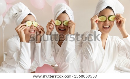 Happy young women wear white bathrobes towels on head make cucumber facial skin care mask on eyes laughing relaxing together, smiling girls friends having fun on spa beauty salon party with balloons