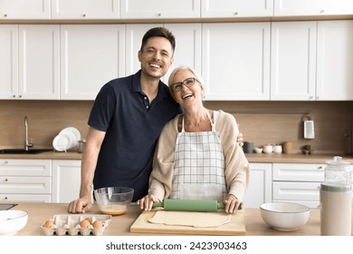 Happy younger man and elder woman cooking homemade bakery food, rolling raw dough for pitta, looking at camera, hugging, smiling, laughing. Happy senior mother and adult son domestic family portrait - Powered by Shutterstock