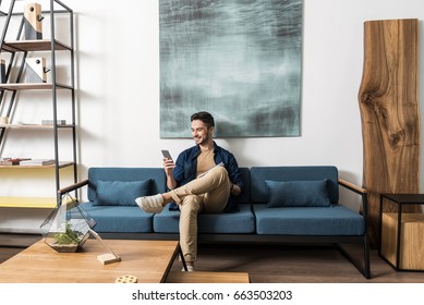 Happy youthful guy bearded resting with cellphone in living room