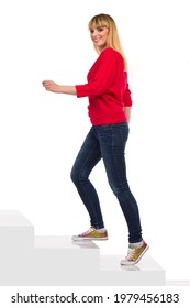 Happy ypung young woman in red sweater, jeans and colorful sneakers is walking up the stairs and looking at camera. Side view. Full length studio shot isolated on white.