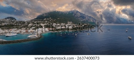 The harbor of Capri Island, Italy, features yachts, a stone jetty, whitewashed hillside buildings, rugged cliffs, and a sunset tinged sky over turquoise waters.