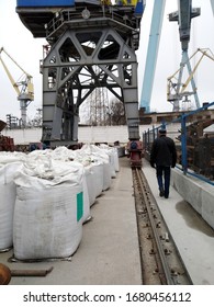 Harbor Crane In A Warehouse. A Man Without A Protective Helmet Goes Along The Warehouse With Big Bags. Violations Of Safety Regulations During Loading And Unloading. Labor Protection At Work