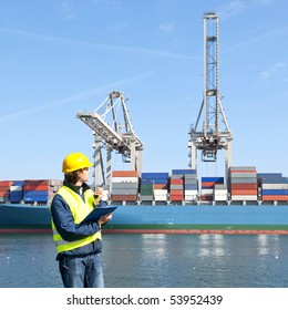 Harbor Dock Worker Talking On His Radio, With A Container Ship Being Unloaded In The Background