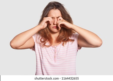 Hard Awakening Concept. Sleepy Young Attractive Woman Rubs Eyes, Has Mess On Head, Awakes Very Early, Dressed In Casual Clothes, Cant Get Up, Isolated Over White Wall In Bedroom. Bed Time And Rest.