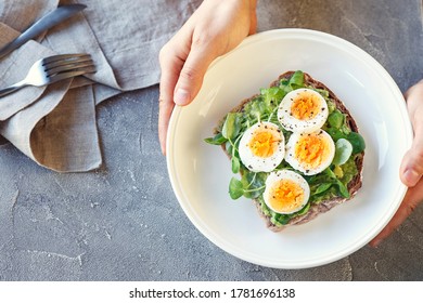 Hard Boiled Egg On Avocado Toast With Green Leaves, Healthy Breakfast Or Lunsh, Top View, Man's Hands Holding A Plate, Grey Background