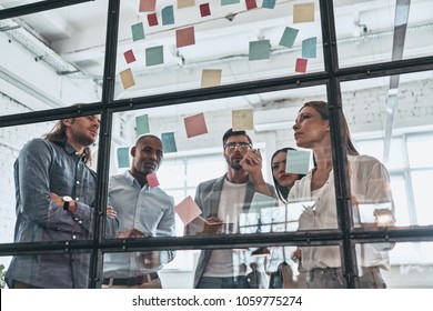 Hard choice to make. Group of young modern people in smart casual wear using adhesive notes while standing behind the glass wall in the board room            - Powered by Shutterstock