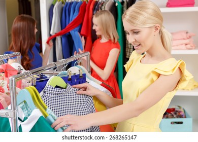 Hard Choice. Young Smiling Blond Woman Looking Through The Clothes On Clothing Rack