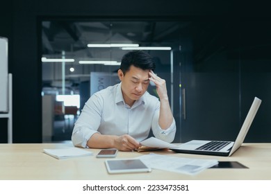 A hard day's work. A serious young Asian man sits tiredly in the office at the desk, works with documents, problems with accounts, holds his head with his hand. - Shutterstock ID 2233473581