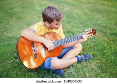 Hard of hearing preteen boy playing guitar outdoor. Child with hearing aids in ears playing music and singing song in park. Hobby art activity for children kids. Authentic childhood moment.  स्टॉक फोटो