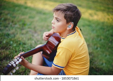 Hard Of Hearing Preteen Boy Playing Guitar Outdoor. Child With Hearing Aids In Ears Playing Music And Singing Song In Park. Hobby Art Activity For Children Kids. Authentic Childhood Moment. 