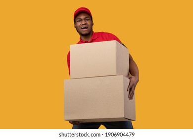 Hard Work. Unhappy black male courier in red cap and uniform holding pile of large heavy cardboard boxes, carrying packages with weight furniture, standing isolated over yellow studio background