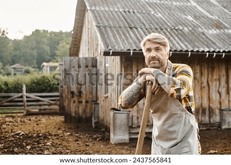hard working appealing farmer with beard and tattoos using pitchfork while working with manure