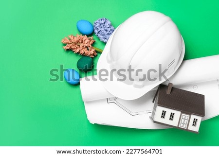 Hardhat with house model, plan, flowers and Easter eggs on green background