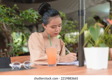 A Hardworking Asian Woman Is Focused On Writing Down Notes While Preparing For Her Upcoming Meeting With A Client In An Al Fresco Cafe.