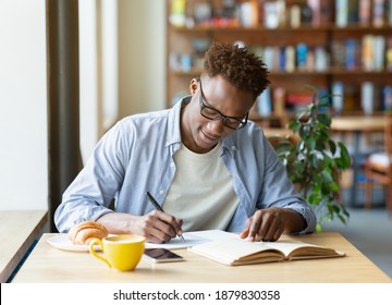 Hardworking black man writing down info from textbook, getting ready for test at cafe. Cool African American guy studying at coffee shop, finding data for his coursework, making home assignment