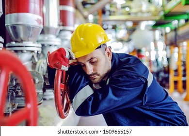 Hardworking Caucasian Worker In Working Clothes Leaning On Big Valve And Sleeping. Night Shift. Energy Plant Interior.