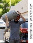 A hardworking man in a dirty shirt hat and sunglasses carries a heavy gas cylinder on his shoulder. The background features a parked car and greenery. Gasman working performs heavy physical labor work