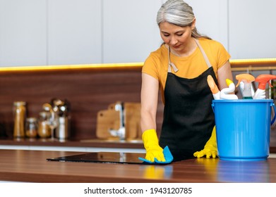 Hardworking mature gray-haired Asian cleaning lady or a housewife in gloves and an apron, stands at kitchen, near a bucket of household chemicals, carefully wipes the stove with rag, cleaning concept