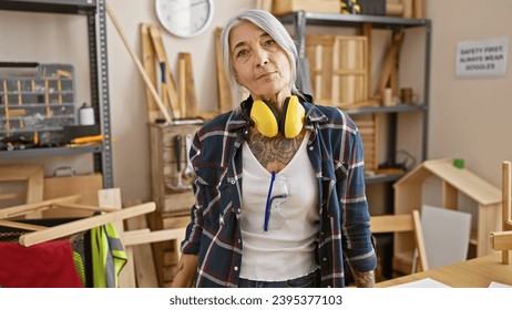 Hardworking middle-aged woman, a grey-haired carpenter with a serious face standing proud in her well-equipped carpentry workshop, headphones on for safety and focus - Powered by Shutterstock