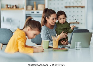 A hard-working mother multitasking by carrying her toddler and working remotely at home on her laptop while her daughter is doing her homework. - Powered by Shutterstock