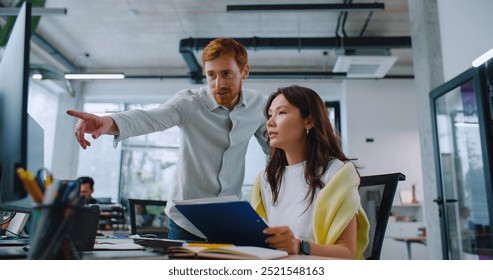 Hardworking multi-ethnic people talking with each other in front of computer. Adorable woman sitting while man standing besides her. People discussing working task or deciding important question.