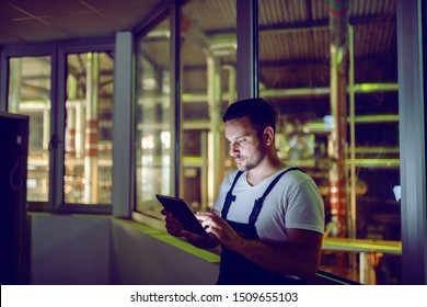 Hardworking Plant Worker In Coveralls Standing And Using Tablet At Night. Night Shift.