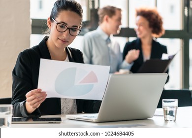 Hard-working young woman analyzing printed and electronic business information while sitting at desk in the office